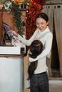Glad adult Asian woman in apron standing near modern cash desk while touching screen and hugging adorable girl while looking at each other during work in cozy cafe