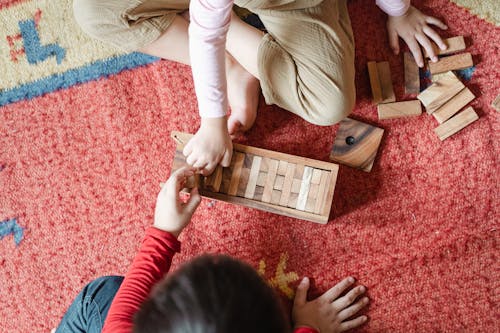 Top view of anonymous barefoot boy and girl in casual clothes sitting on floor carpet and playing with wooden blocks of jenga tower game