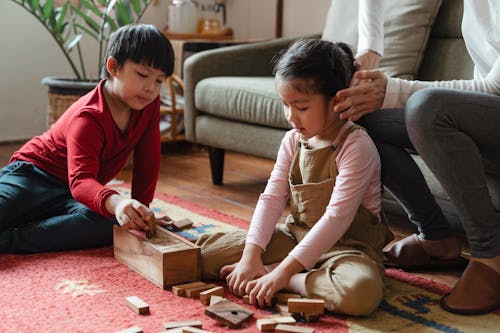 Free Unrecognizable mother sitting on sofa in modern living room while spending time together with Asian son and daughter sitting on floor carpet and playing jenga game Stock Photo