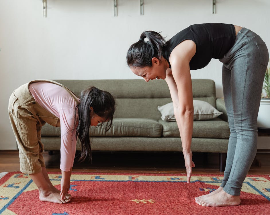 Free Side view of smiling young Asian woman stretching body with adorable daughter while standing against each other on floor carpet near cozy couch in modern living room in morning Stock Photo
