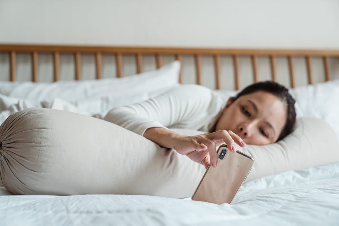 Content young ethnic female watching video on cellphone while lying on comfortable bed with long cylinder pillow in morning and enjoying free time