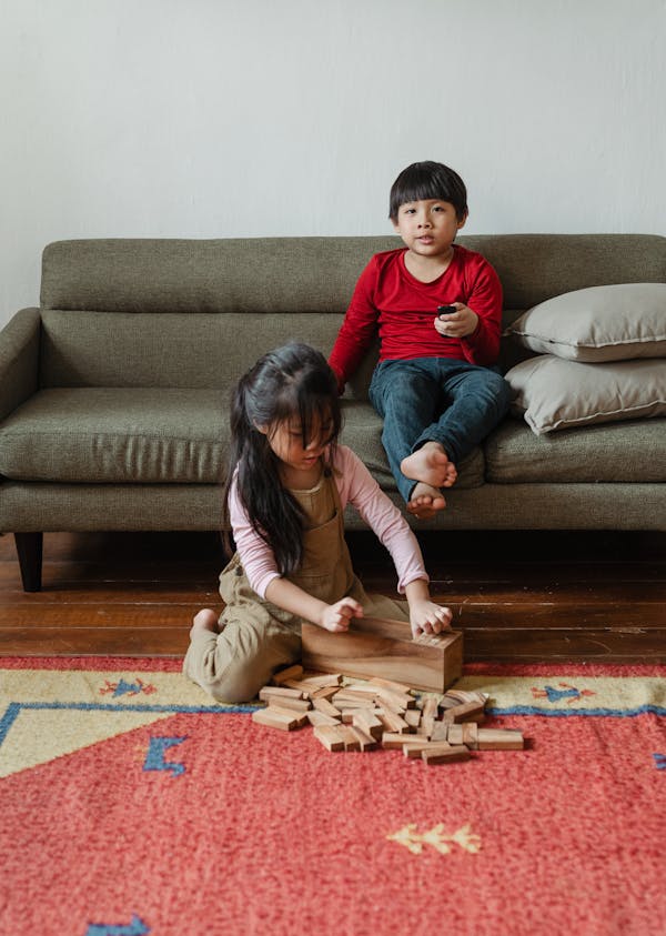 Barefoot ethnic boy sitting on cozy sofa and switching TV channels with remote control while cute sister playing wooden tower game sitting near on floor carpet at home