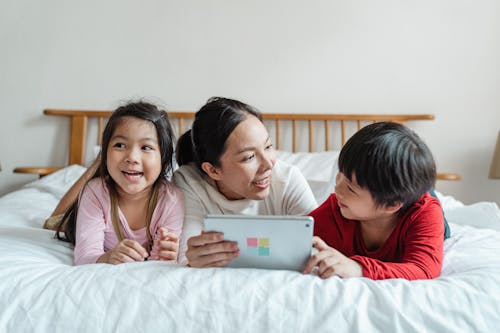 Full length smiling ethnic mother lying on stomach on comfortable bed with little daughter and son while using tablet in light room