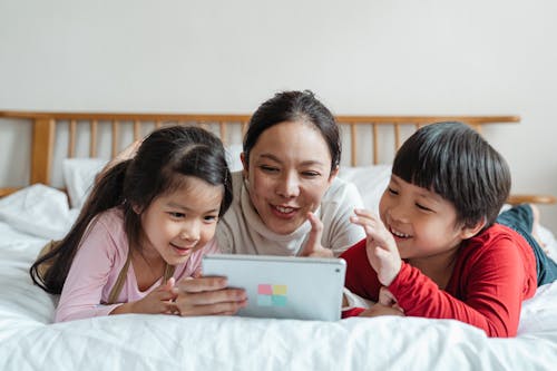 Free Full body smiling Asian mother and kids in casual clothes lying on stomachs on comfortable bed while spending time together playing game via tablet Stock Photo