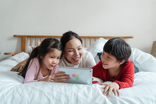 Cheerful ethnic mother watching video via tablet with kids on bed