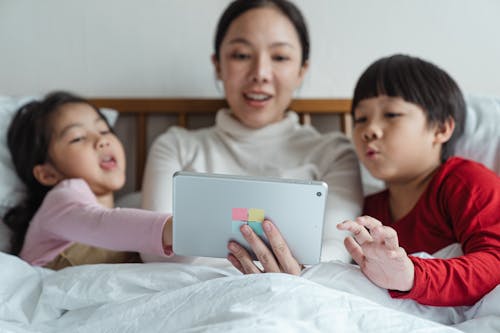 Interested Asian mother and little daughter and son in pajamas lying in comfortable bed choosing content in Internet on tablet while spending time together in morning