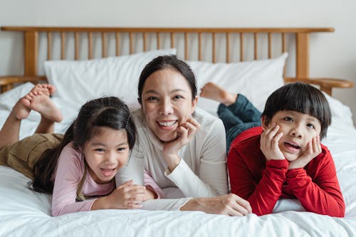 Free Cheerful Asian mother with children in casual clothes lying on stomach leaning head on hand having rest on bed together Stock Photo