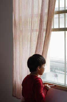 室内，一个年幼的孩子挂着透明窗帘，若有所思地看着窗外