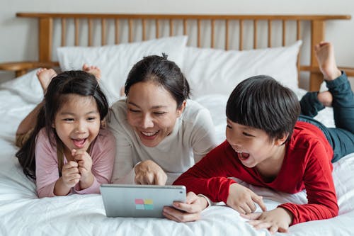 Full body happy Asian mother pointing at screen tablet showing interesting information to cheerful kids while lying on cozy bed in light room