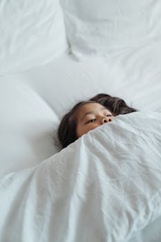 一个孩子安静地躺在床上，盖着一条白色的毯子，营造出舒适轻松的早晨氛围