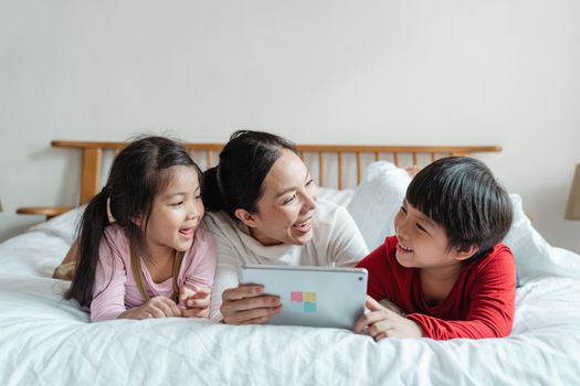 在明亮的公寓里，一位快乐的亚洲女性和一个微笑的小孩子穿着休闲服，躺在柔软的床上使用平板电脑