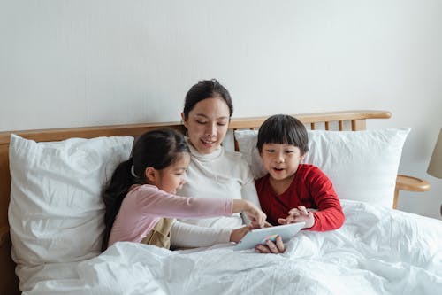 Photo of Woman and Kids Sitting on Bed While Using Tablet Computer