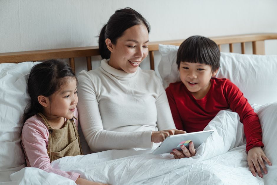 Asiatische Mutter und Kinder schauen sich Tablet an und fühlen sich im Bett verbunden.