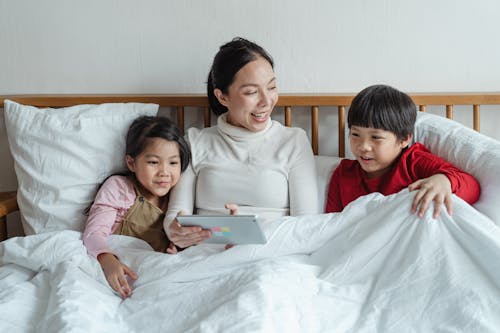 Free Positive Asian mother and little daughter and son in pajamas resting together under soft blanket in bed while using tablet and watching interesting video in morning Stock Photo