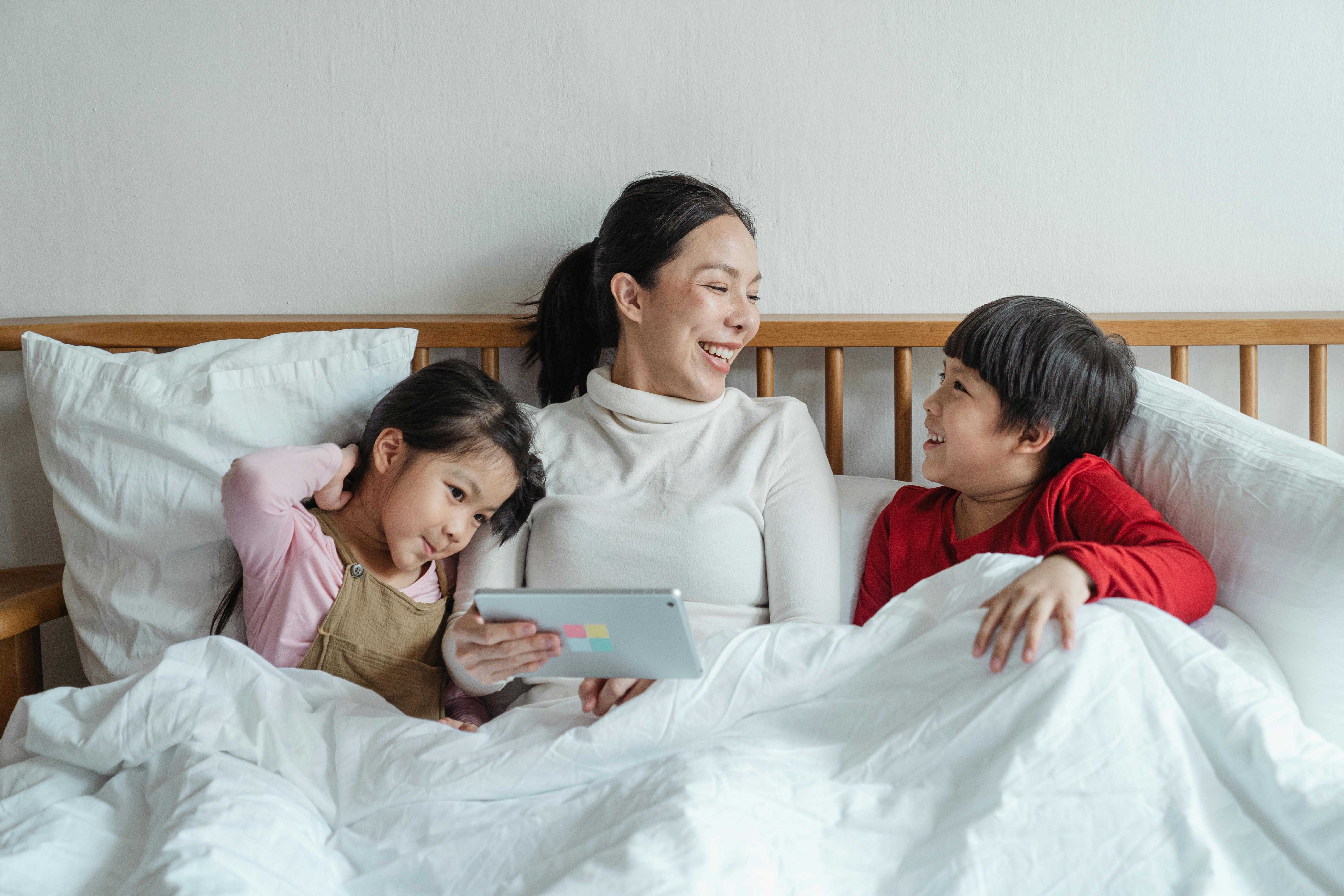 cheerful asian mother and little children with tablet having fun in bed