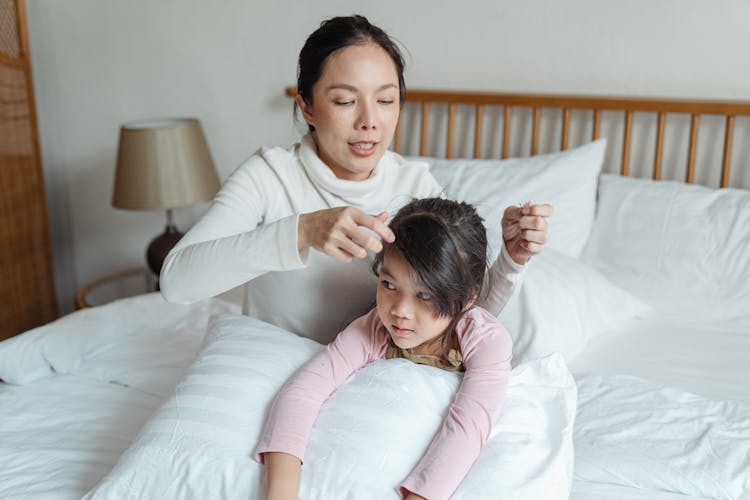 Asian Mother Adjusting Hair Of Daughter On Bed