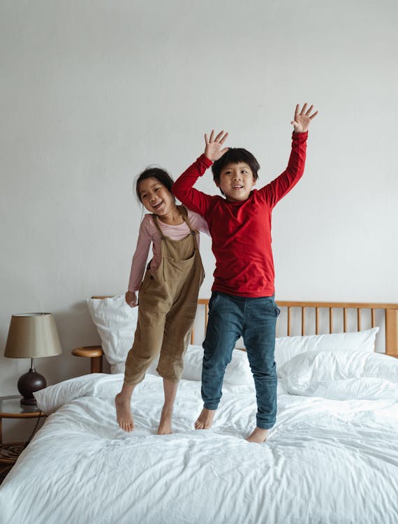 Full body cheerful Asian children wearing casual clothing standing with arms raised on comfy bed and smiling while playing together