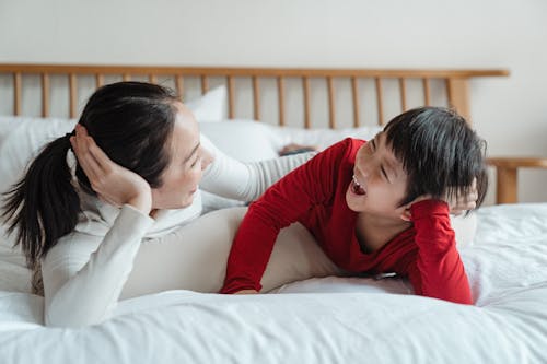 Full length delighted happy Asian woman with cute son resting on cozy soft bed and laughing while spending morning together at home