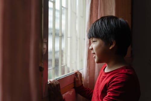 Side view content little Asian boy in red shirt wrinkling nose and grimacing while standing near window and looking out