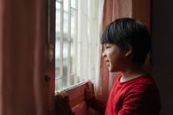 Side view content little Asian boy in red shirt wrinkling nose and grimacing while standing near window and looking out