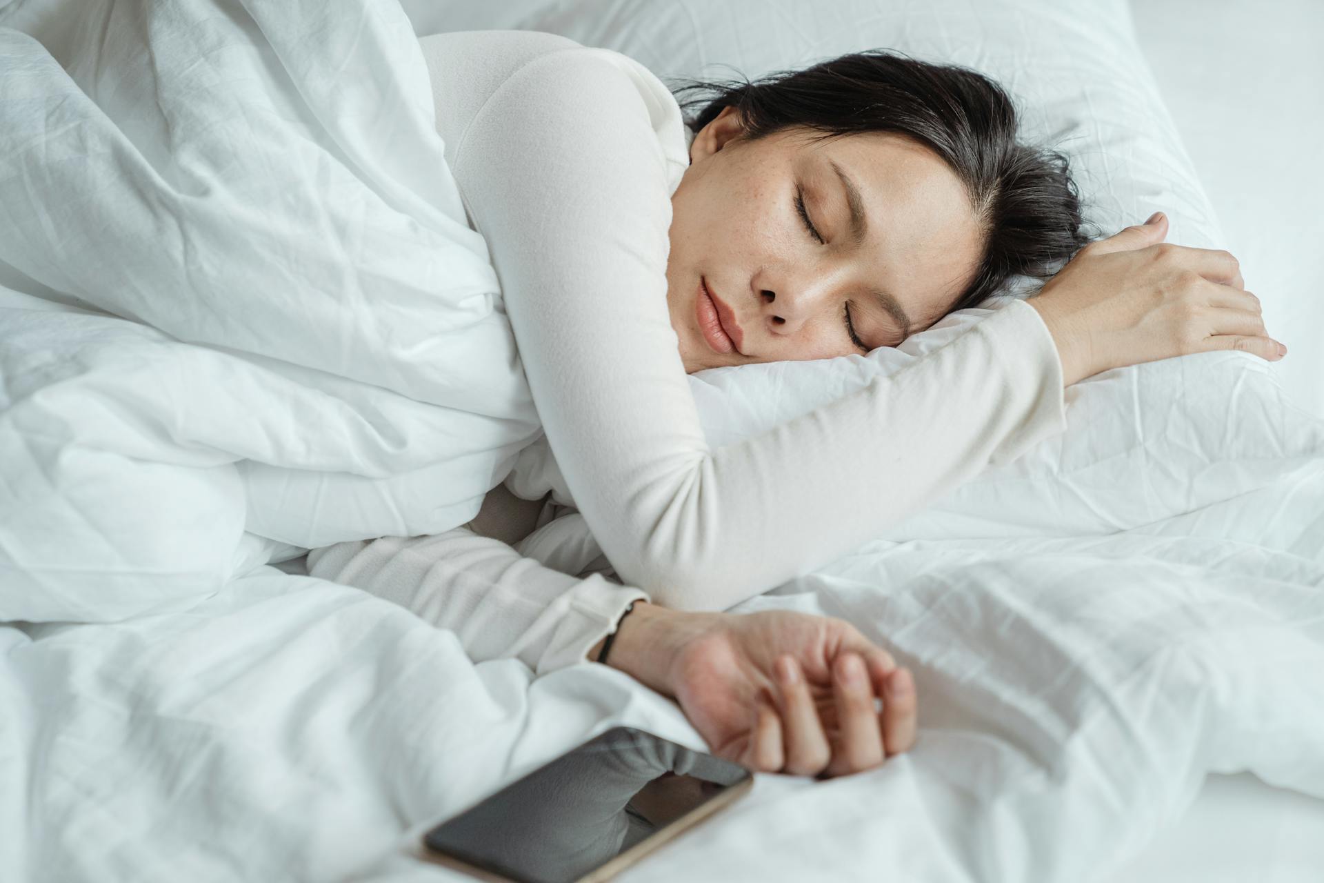 Calm Asian female wearing white pajama sleeping in comfortable bed with white sheets near modern mobile phone in morning