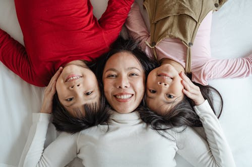 Free Top view of excited smiling Asian mother caressing adorable little kids in bright casual clothes while resting together on comfortable bed with white sheets and looking at camera Stock Photo