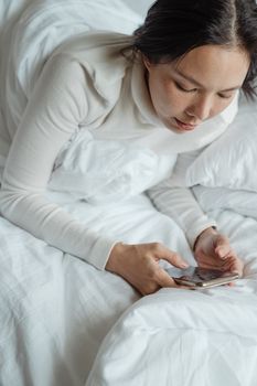 A woman in a white sweater uses her smartphone while relaxing in bed, enjoying leisure time indoors.