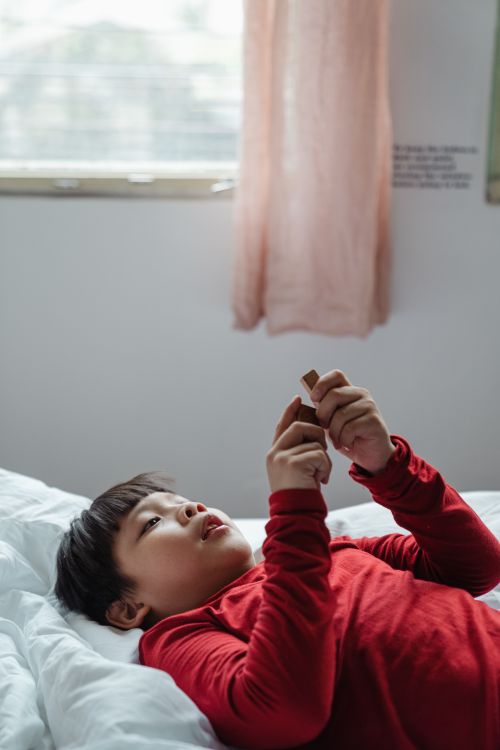 Free Glad cute Asian boy in red pajama lying on bed near window and playing in sunny morning Stock Photo