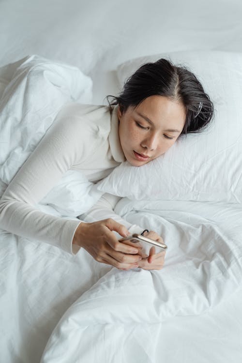 Free From above calm young Asian female in sleepwear waking up in morning and browsing mobile phone in cozy bed Stock Photo