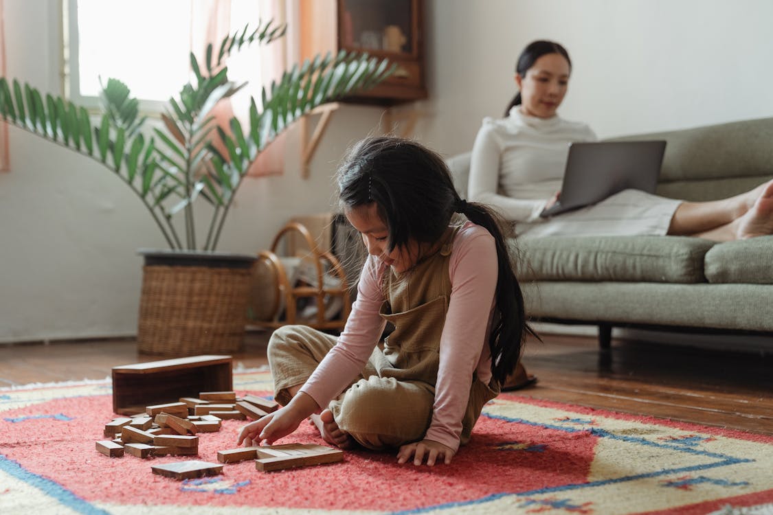 Content little Asian girl in casual wear sitting on carpet on floor and playing tower game while focused mother working on netbook on sofa near houseplant