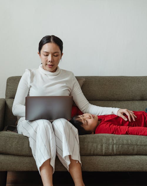 Positive Asian woman working on laptop on sofa in living room and hugging calm adorable son in casual clothes