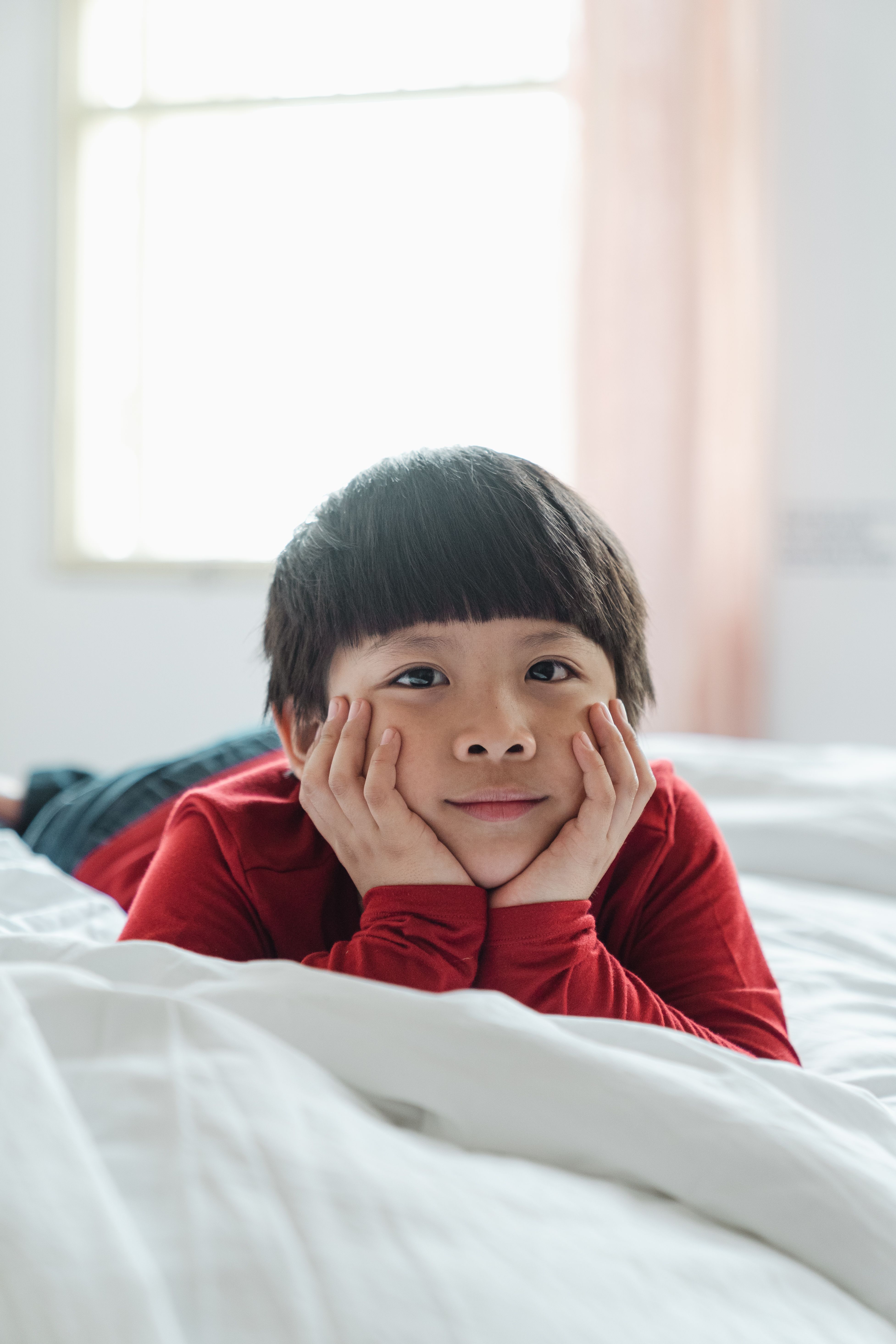 一个快乐的孩子穿着红衬衫，在阳光明媚、舒适的卧室床上放松身心，享受着宁静的早晨