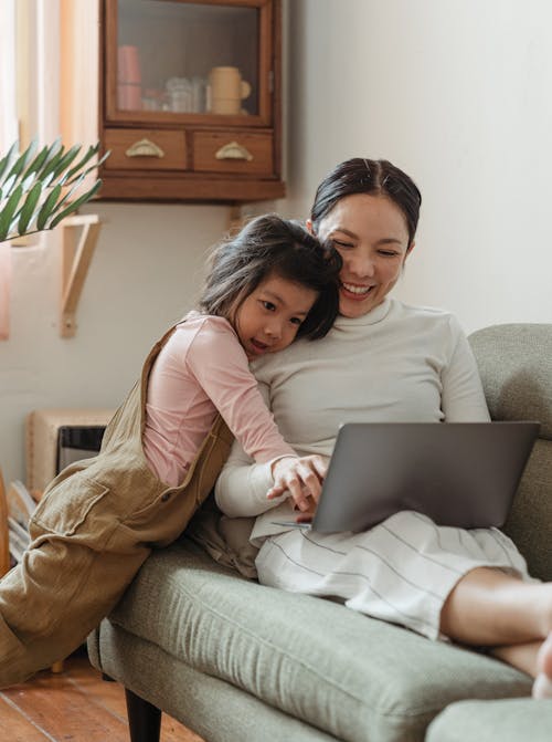 Cheerful smiling Asian woman browsing modern netbook while hugging with cute content daughter on comfy sofa in cozy living room