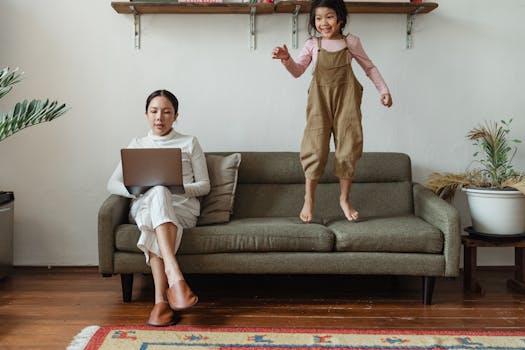 Mother works on laptop at home while child jumps on sofa, showing modern family life.