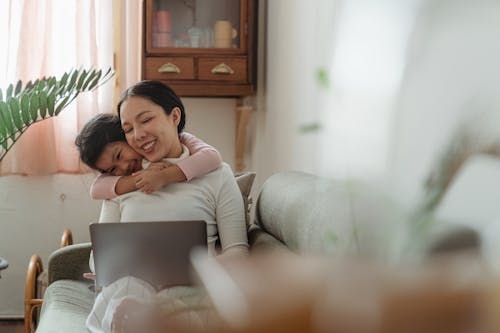 Free Excited cute Asian daughter bonding with smiling happy mother using netbook on cozy sofa in living room in daytime Stock Photo