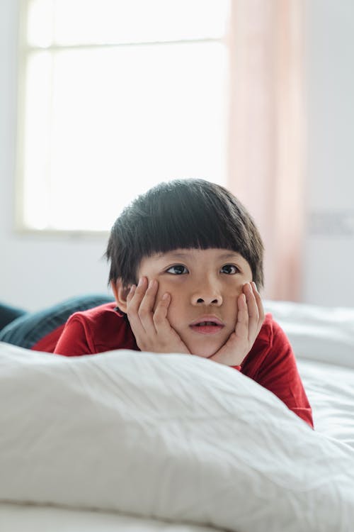 Amazed cute Asian boy lying on cozy bed against bright window and holding face between hands while looking away surprisingly