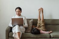 一位母亲在笔记本电脑前工作，她的女儿则躺在沙发上，双腿抬起，营造出舒适的家庭氛围