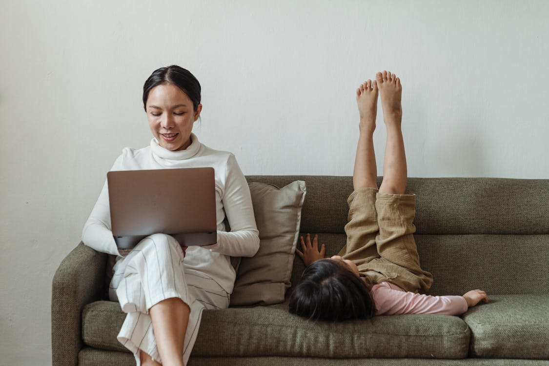 Free Cheerful mother working on laptop near daughter on couch Stock Photo