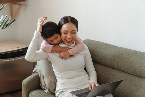 Free Positive smiling Asian girl in casual wear embracing happy mother working on laptop on cozy couch in daylight Stock Photo