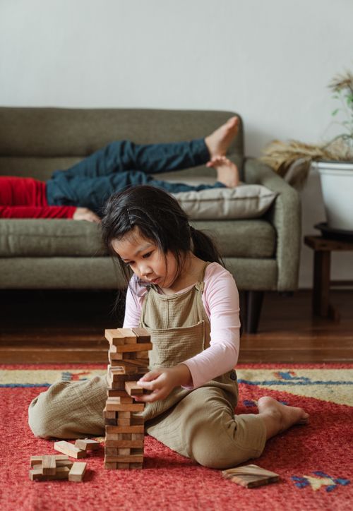 Free Adorable content Asian girl in casual clothes sitting on carpet on floor and playing tower stacking game near brother resting on sofa Stock Photo