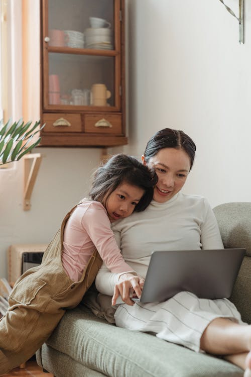 Free Positive smiling Asian woman in white casual clothes resting on cozy sofa and browsing netbook with cute little daughter Stock Photo