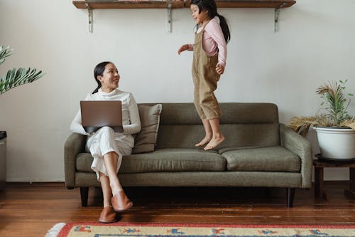 Free Smiling Asian mother in casual wear working on laptop while cute daughter jumping on couch in cozy living room Stock Photo