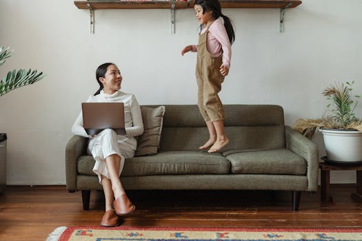Smiling Asian mother in casual wear working on laptop while cute daughter jumping on couch in cozy living room