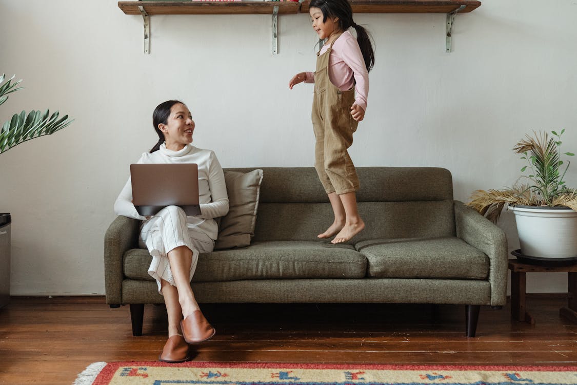 Smiling Asian mother in casual wear working on laptop while cute daughter jumping on couch in cozy living room