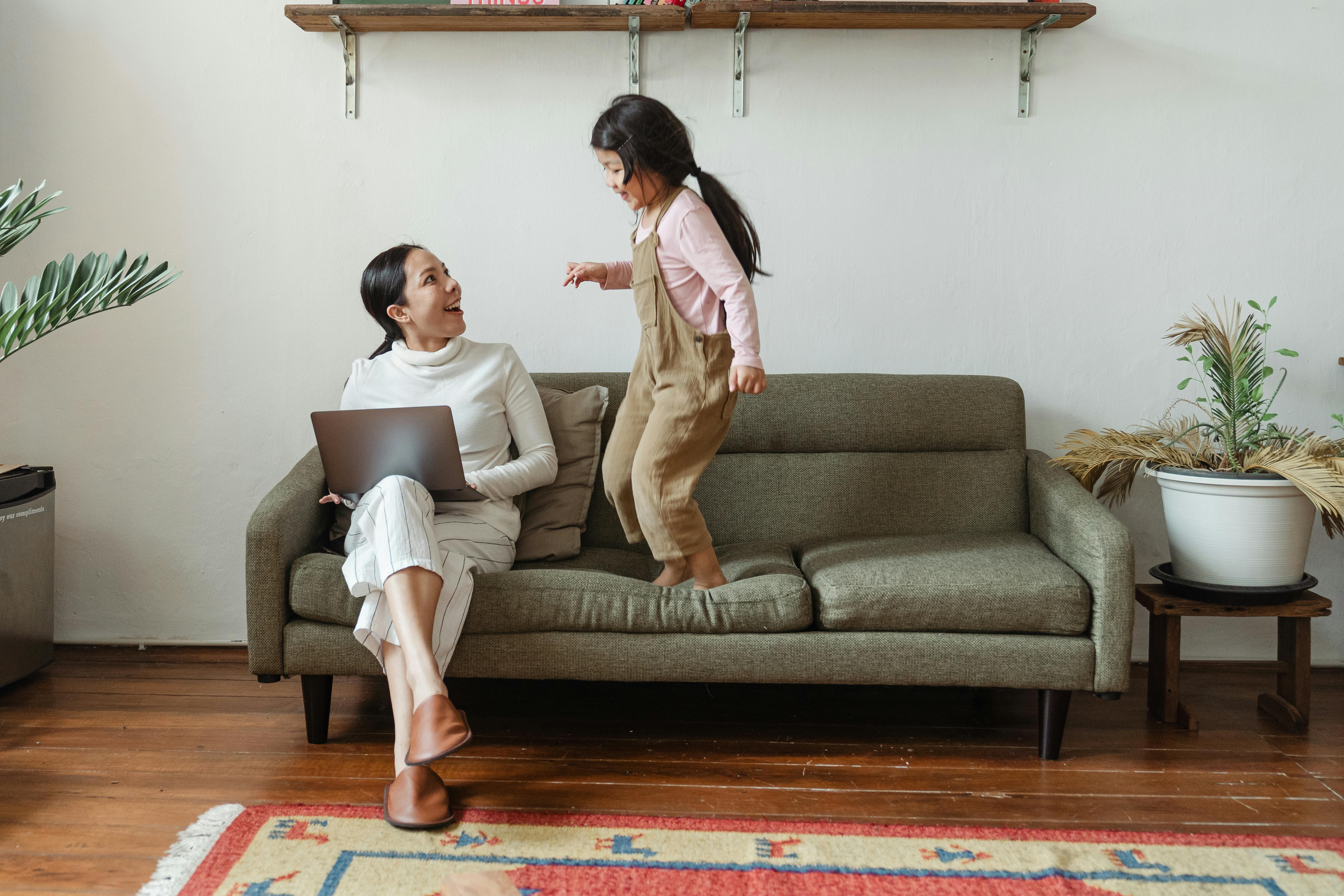 happy mother with laptop talking to little daughter on couch