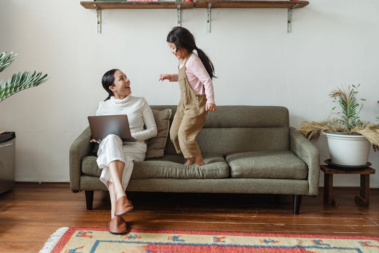 Happy Mother With Laptop Talking To Little Daughter On Couch