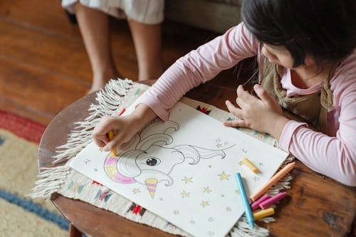 From above crop anonymous little girl in casual wear coloring unicorn in coloring book while sitting at table near sibling