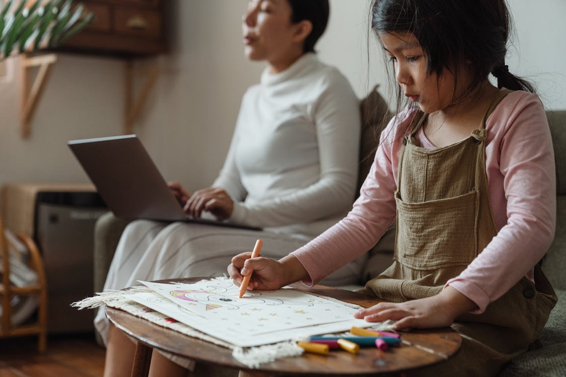 Free Focused girl drawing on paper near mother with laptop Stock Photo