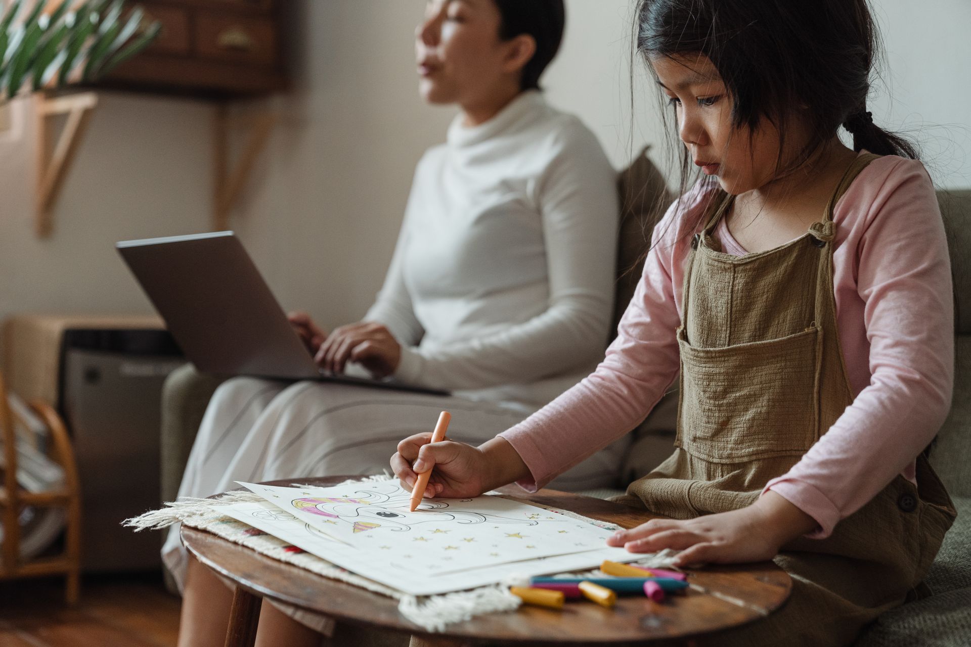 A mother works on a laptop while her daughter colors in a cozy living room, balancing work and family life.