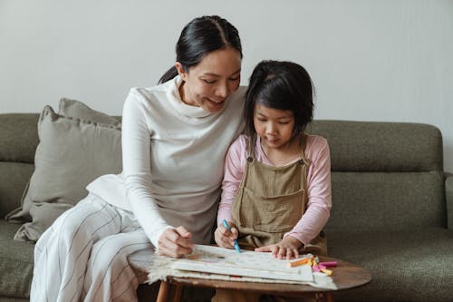 Happy smiling Asian mother and cute little girl sitting on sofa and coloring picture with multicolored crayons